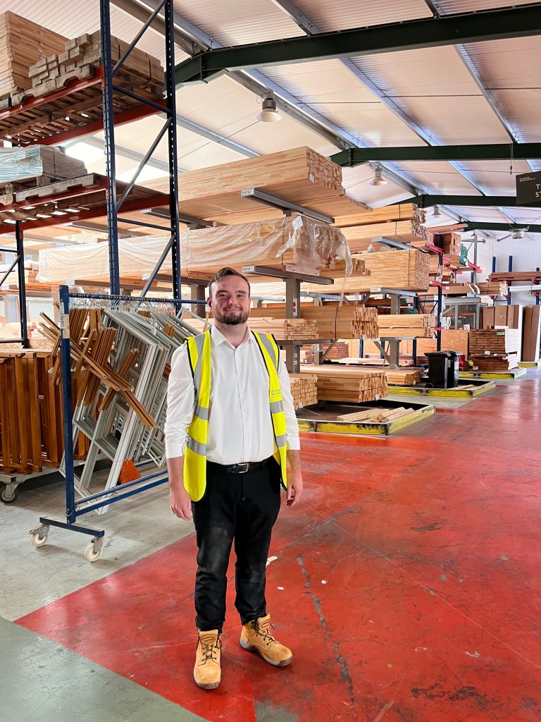 Daniel folkes standing in a timber warehouse with shelves of long pieces of timber behind him