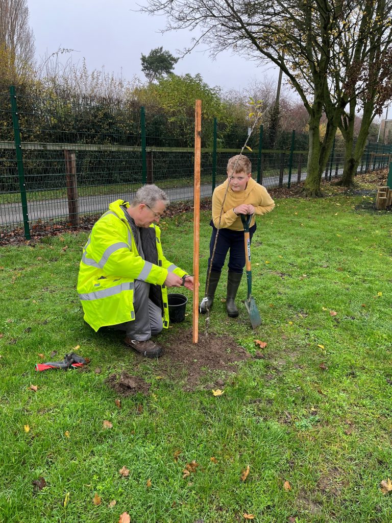 Factory operative assisting the only boy in the group to plant a tree