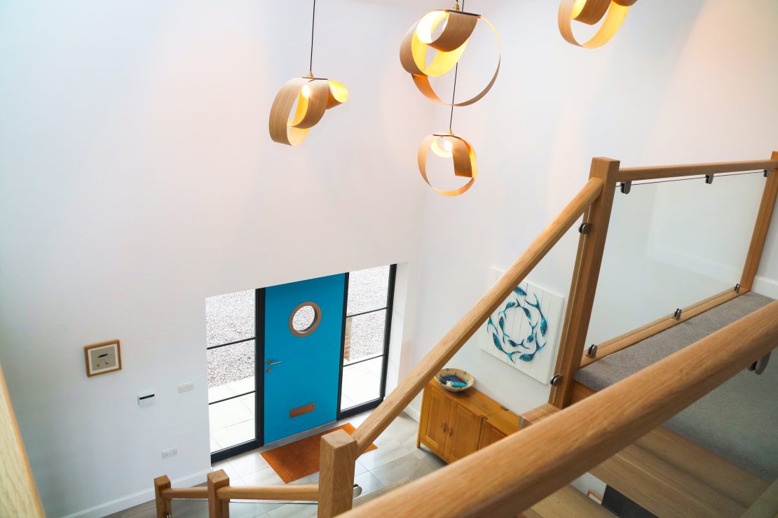 Blue front door with a stainless steel framed porthole window and anthracite grey framework offset by bespoke timber staircase